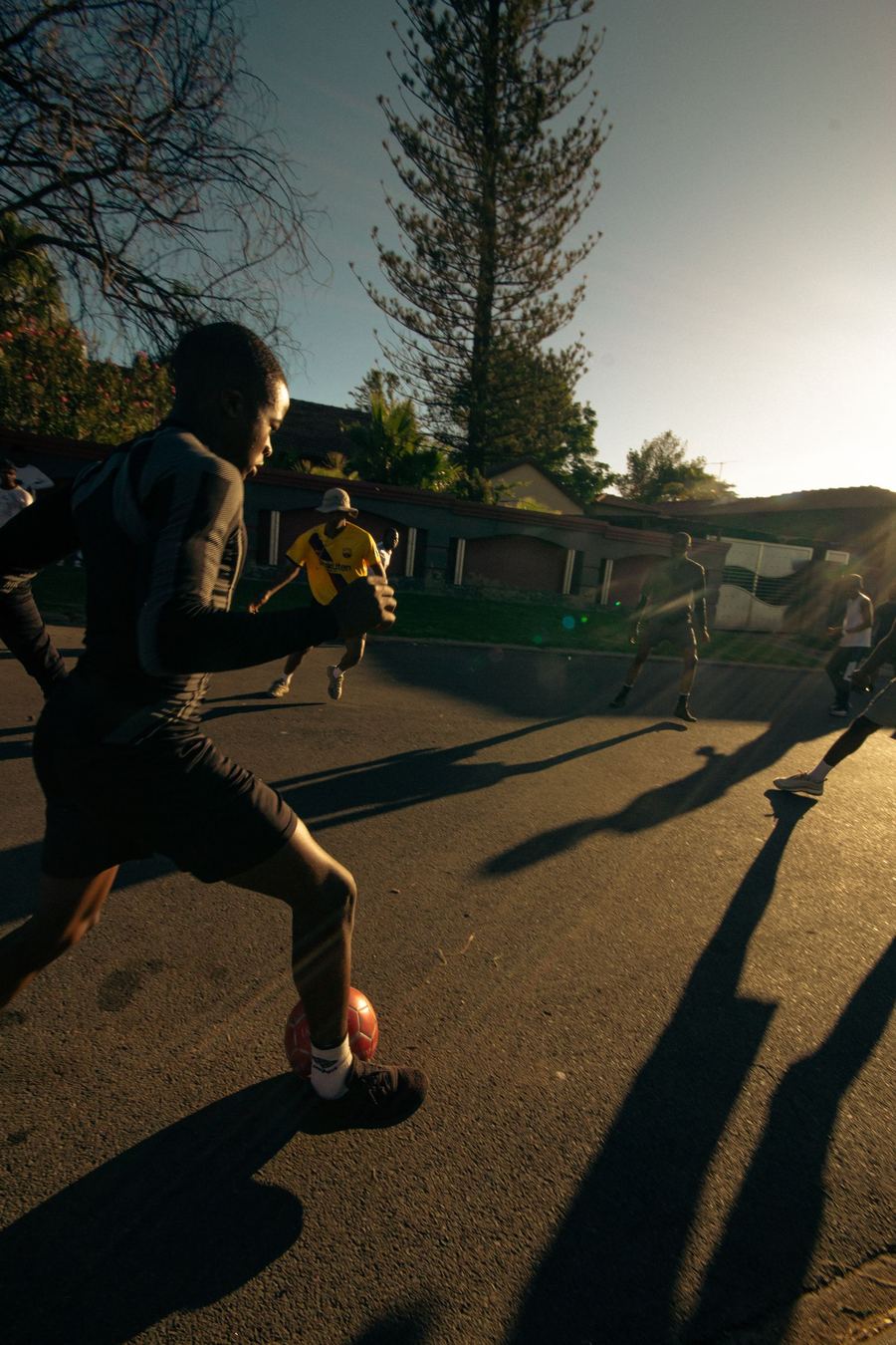 A street football game at golden hour, a player sprinting with the ball as long shadows stretch across the road